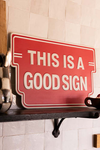 Red sign with white text 'THIS IS A GOOD SIGN' on a wooden shelf.