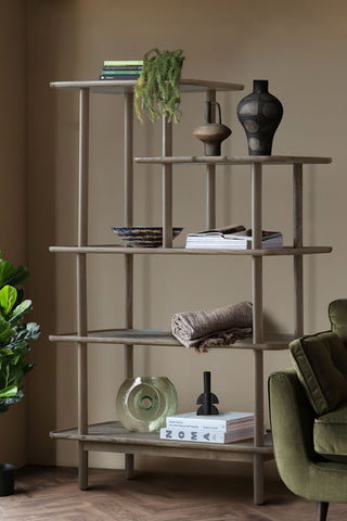 The Tiered Oak Display Cabinet In Grey styled in front of a neutral wall with various home accessories on, styled next to a green sofa and a plant.