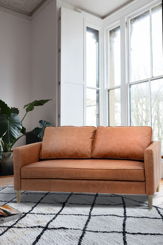 The Vintage-Style Brown Leather 2-Seater Sofa styled on a rug in a living room, with a large window and plant in the background.