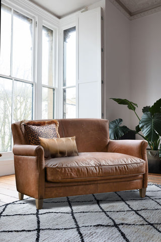 The Vintage-Style Dark Brown Leather 2-Seater Sofa styled with cushions on, on a rug in a living room, with a large window and plant in the background.