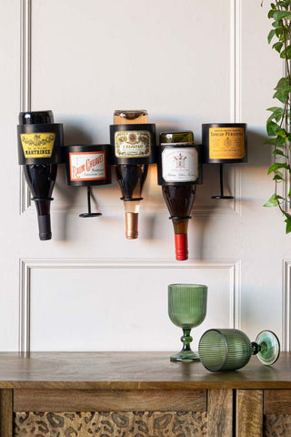 The Vintage-Style French Wall-Mounted Bottle Holder with bottles inside, styled on the wall above  sideboard and some glasses, next to a plant.