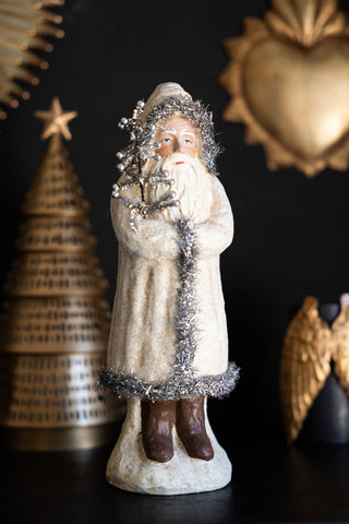 Decorative figurine of a bearded man in a long coat with tinsel, against a dark background with festive decorations.