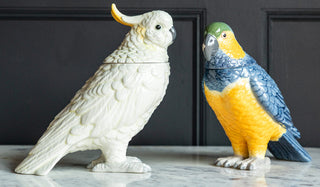 The Ceramic Cockatoo Storage Jar and Ceramic Parrot Storage Jar displayed together on a tabletop in front of a black panelled wall.