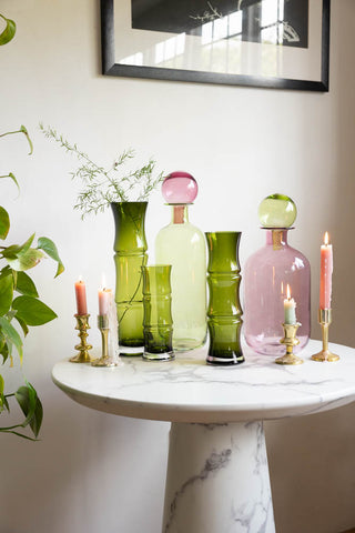 The Green Glass Bamboo Vases in each size displayed with the Small & Large Green and Pink Apothecary Bottles on a white marble table with lit candles. There is a plant and art print in the background. 