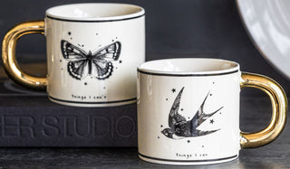 The Monochrome Set of 2 Tattoo Mugs displayed on a black table with a book.