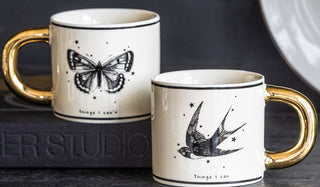 The Monochrome Set of 2 Tattoo Mugs displayed together on a black sideboard and book, with a plate in the background.