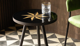 The Small Black Star Side Table with a glass on top, displayed on a geometric floor next to a green chair in front of a dark wooden wall.