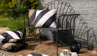 Garden lifestyle image of the Black & Natural Stripe Outdoor Cushion and Black & Green Stripe Outdoor Cushion displayed together on and next to a bench in the sunshine, with magazines, a mug and a jug to the side of the shot.