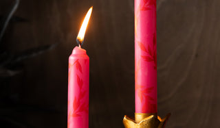 Two of the Set Of 4 Pink Leaf Dinner Candles displayed lit in a gold candlestick holder in front of a dark wooden wall.