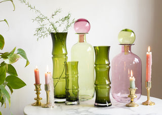Lifestyle image of the Green Glass Bamboo Vases with the Small & Large Green and Pink Apothecary Bottles displayed together on a marble table with lit candles. There is a plant and art print displayed in the background of the shot. 