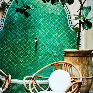 Square image of the green courtyard garden. in the background is a green tiled outdoor bathroom. In the foreground, potted trees and wicker chairs.