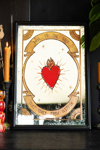 A decorative rectangular mirror with the words, 'Love Is The Drug' on it, plus a red milagro-style heart. Styled on a black surface with candlestick holders and greenery.