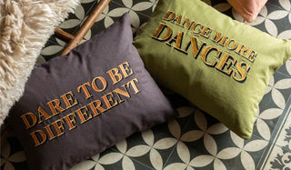 Lifestyle image of the Dance More Dances Embroidered Green Cushion Cover and the Dare To Be Different Embroidered Brown Cushion Cover displayed together on a geometric floor next to a wicker bench and sheepskin rug. 