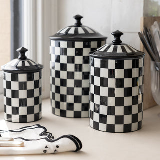 The Set Of 3 Enamel Black & White Check Tins displayed together on a kitchen worktop next to a window, with a napkin, chopping board and a glass filled with cutlery.