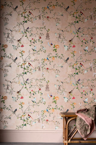 Image of a pink printed wallpaper with birds, branches and flowers, displayed on a wall behind a wooden bench with a throw on top. 