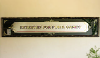 The Reserved For Fun & Games Vintage-Style Mirror on a neutral wall with a plant in the corner of the shot.