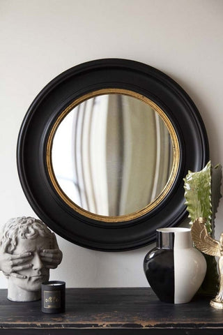 A round wall mirror with a wooden frame styled above a shelf with ornaments, a vase and a candle