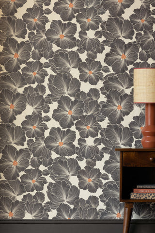 Black, white and orange floral repeat printed wallpaper, displayed on a wall behind a small table with lamp. 