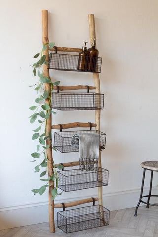 Lifestyle image of the Wooden Ladder With 5 Basket Shelves with green vine leaves wrapped around the ladder and the baskets filled with soap bottles and towels.