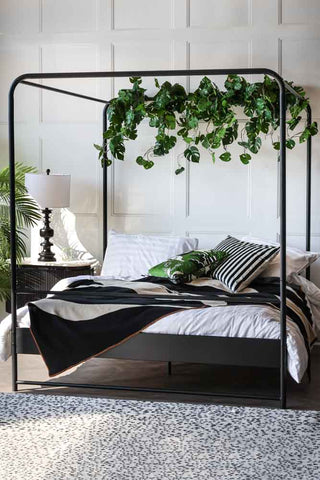 An image of a black metal four-poster bedframe, styled with greenery and luxurious bed linen, pillows and cushions 