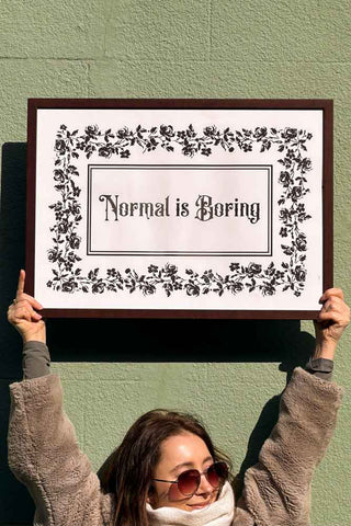 A woman holding up an art print that says, 'Normal Is Boring'