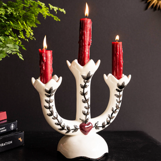 A colourful gold candlestick holder with two love bird figures on, styled with some lit candles on a table with a plate and a cocktail glass.