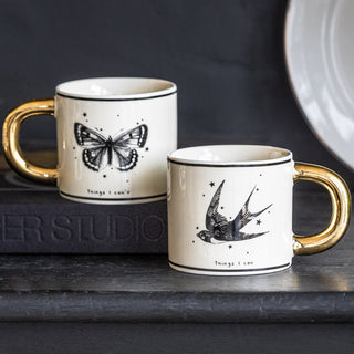 The Monochrome Set of 2 Tattoo Mugs displayed together on a black sideboard, with a book and plate.