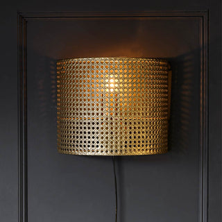 The Gold Metal Wicker Effect Plug In Wall Light displayed illuminated on a black panelled wall.