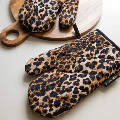 A pair of leopard print oven mitts styled on a white surface with a wooden chopping board.