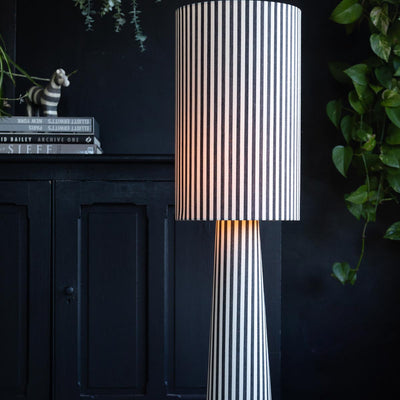 A black and white stripe floor lamp in front of a dark background with greenery.