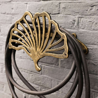 The Pretty Antique Brass Hose Holder displayed on a black brick wall with a hose wrapped around it.