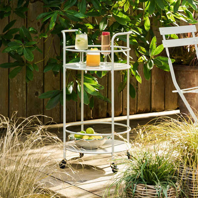 A drinks trolley styled outdoors with various items on, displayed on a patio amongst plants and a dining table.