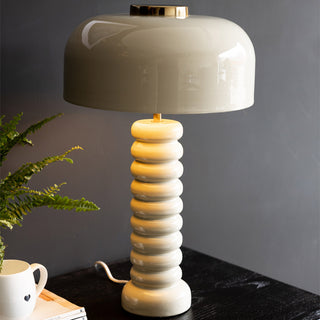 Lifestyle image of the Mushroom Table Light illuminated and displayed on a striped side table, next to a leopard print chair and green velvet curtains. 