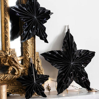 Lifestyle image of the Set Of 3 Black Gloss Paper Stars displayed alongside a mirror, fairy lights and candle on a shelf.