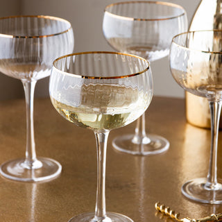 Lifestyle image of the Set Of 4 Ribbed Champagne Coupe Glasses With Gold Rim displayed on a gold tabletop alongside a corkscrew and a gold bottle. 