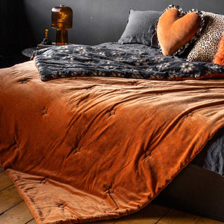 Rockett St George Burnt Orange Reversible Velvet Throw on a bed, styled with a glass amber table lamp and cushions in front of a dark wall. 