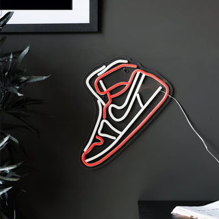 Lifestyle image of the High Top Sneaker Neon Wall Light displayed on a black wall, with a side table, plant and art print in the shot.