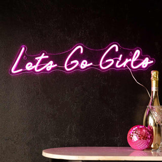 The Let's Go Girls Neon Wall Light on a dark wall above a table styled with a pink disco ball, gold bottle and martini glass.