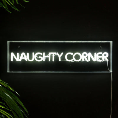 A neon sign that says 'naughty corner' styled on a black wall with some greenery.
