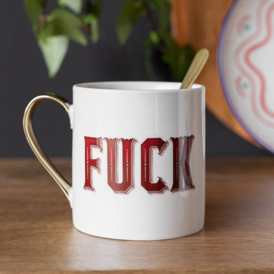 A white and gold mug with red text reading 'fuck' displayed on a wooden surface witha plant and some plates in the background.