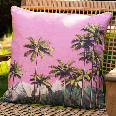 A pink pal tree print cushion styled on a wicker seat outdoors.