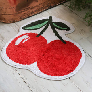 The Cherries Bath Mat displayed on a wooden bathroom floor in front of a copper bath and plant.
