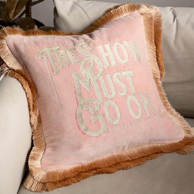 A pink fringed cushion with the words 'the show must go on', displayed on a neutral sofa.
