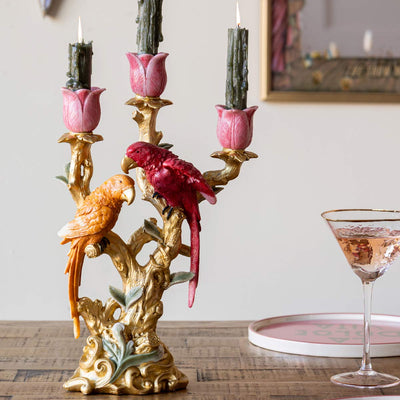 A gold candle holder with two decorative lovebirds on, styled on a wooden table with a cocktail glass and a plate.