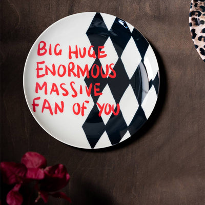 A decorative wall plate that reads 'big huge enormous massive fan of you' displayed on a dark wall next to another plate and some flowers.