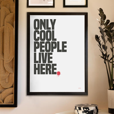 An art print reading 'only cool people live here' displayed on the wall with other prints, styled with a potted plant and trinket box on a table beneath.