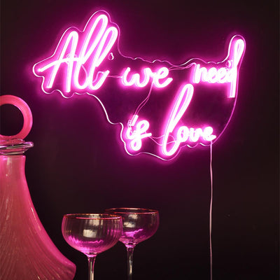 A pink neon sign reading 'all we need is love', styled on the wall above a pink decanter and some cocktail glasses.
