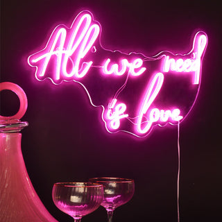 Lifestyle image of the Rockett St George All We Need Is Love LED Acrylic Neon Light displayed illuminated on a dark wall and styled together with a glass decanter and two champagne glasses. 