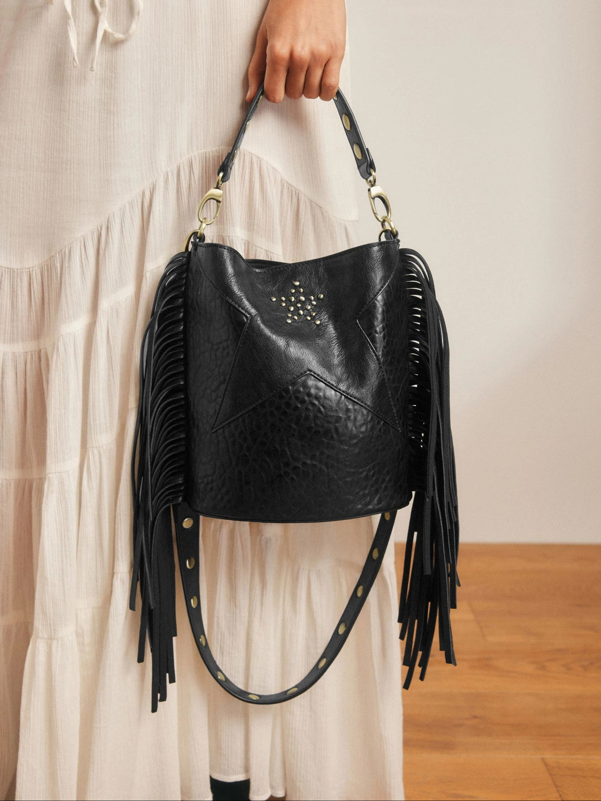 Black leather handbag with fringe details held by a person wearing a light-colored dress.