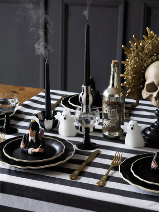 A styled halloween table with black and white stripe tablecloths, halloween decorations such as ghost candles, a wednesday ornament and black and white tableware.